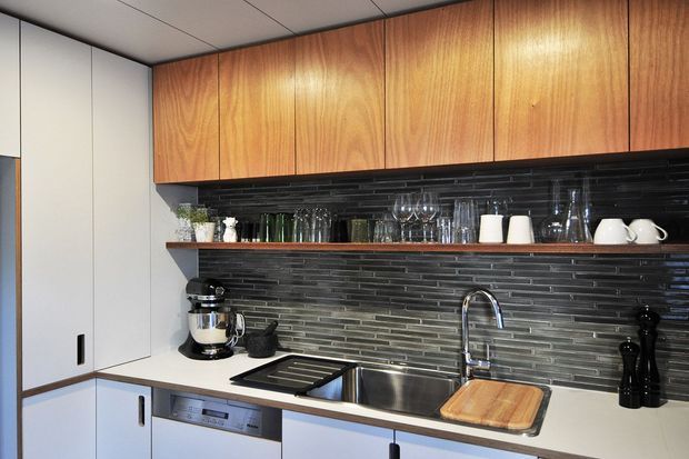 Compact contemporary kitchen with flat-panel white lower cabinetry, warm timber overhead cupboards, open shelving, a dark stacked-tile splashback, stainless-steel sink and tap, integrated dishwasher, and a light benchtop. Glassware and small appliances are arranged on the open shelves and counter.