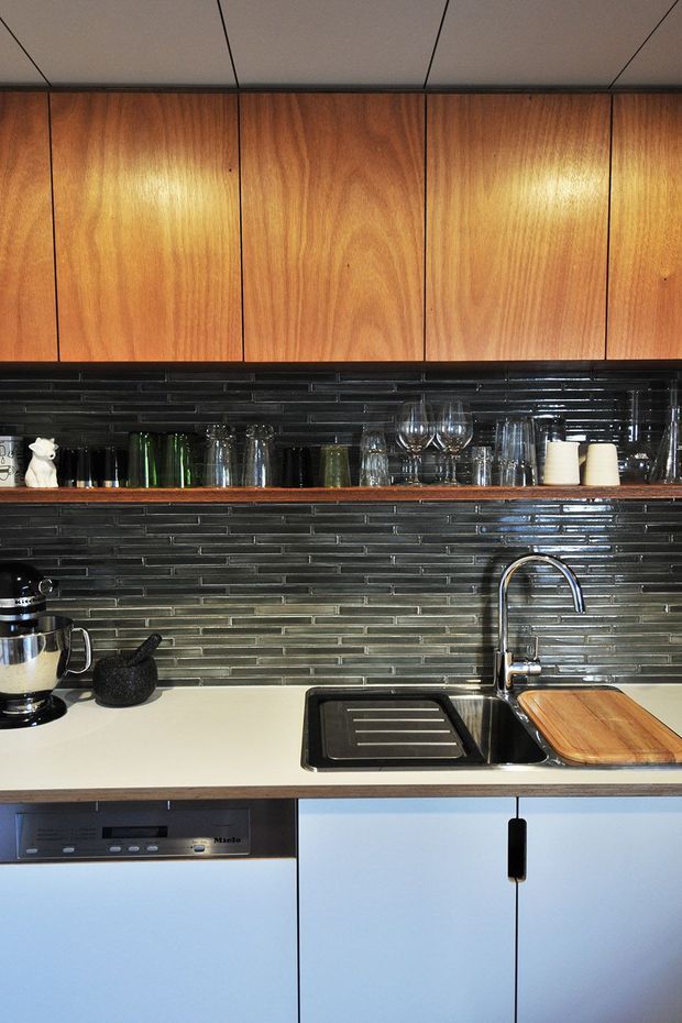 Compact contemporary kitchen viewed straight on, featuring warm timber overhead cabinetry, glossy dark narrow-format tiled splashback, open display shelving with glassware, a pale solid benchtop, stainless-steel sink and tap, integrated dishwasher, and pale blue lower cabinetry. The scheme combines warm wood with dark reflective tile and cool cabinetry.