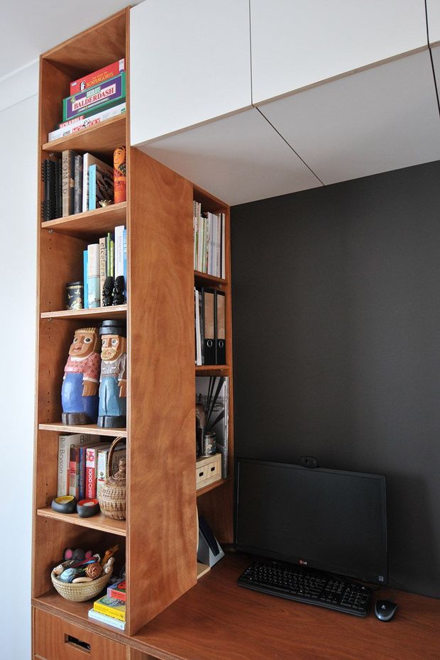 Compact built-in home workspace integrated into a tall timber shelving unit. Warm wood cabinetry and open shelves display books and decorative objects, while a dark charcoal wall frames a computer monitor and keyboard on the desk surface. White overhead cabinets and a clean, tailored fit-out create a highly organized study nook.