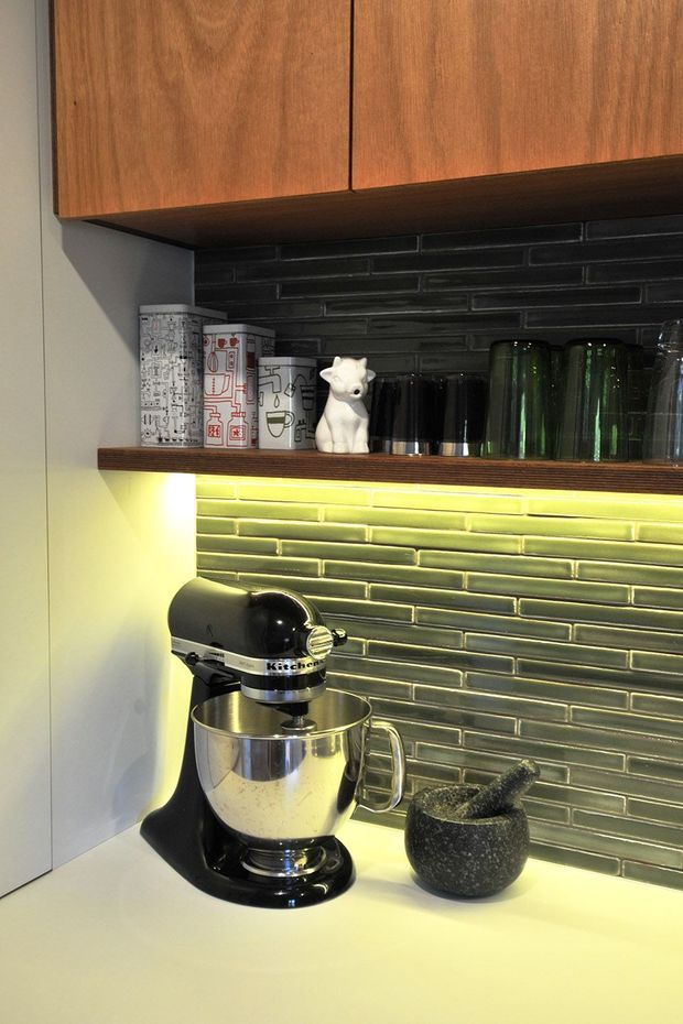 Close-up view of a compact kitchen work area with timber overhead cabinetry, a dark glossy brick-pattern tiled splashback, and a pale benchtop. A black stand mixer and dark stone mortar and pestle sit on the counter, with glassware and patterned canisters arranged on an open shelf. Warm under-cabinet lighting highlights the green-grey tile surface.
