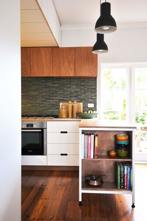 Bright compact kitchen featuring warm timber overhead cabinetry, a dark narrow-format tiled splashback, white lower cabinets, a built-in oven and cooktop, and a timber island or open shelving unit. The space has polished dark timber flooring, large white-framed windows, and two black pendant lights. Open shelving displays books and colourful crockery.