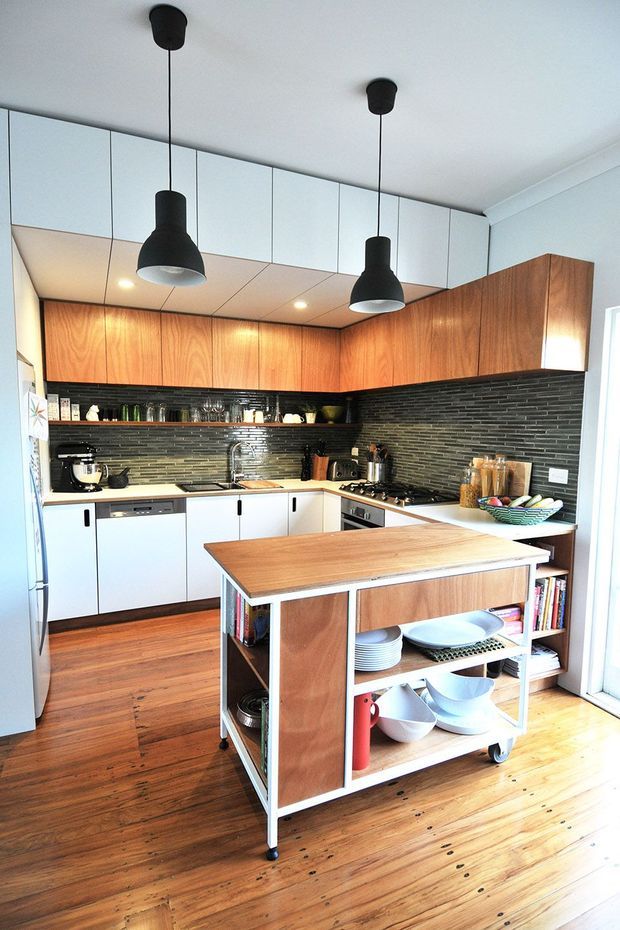 Bright, compact kitchen with a central mobile island on casters. The room features warm timber flooring and upper cabinetry, white lower cabinetry, a dark horizontally tiled splashback, open shelving, stainless-steel appliances, and two black pendant lights. The island has a timber benchtop, white framing, and open storage for dishes and kitchenware.