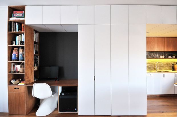 A compact contemporary built-in study and storage wall integrated into an open-plan kitchen. Floor-to-ceiling white flat-panel cabinetry spans the centre, with warm timber shelving, a dark recessed desk nook, books, and a white sculptural desk chair on the left. The kitchen is visible through the opening on the right, featuring timber cabinetry, illuminated shelving, white lower cabinets, and timber flooring.