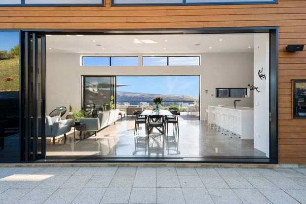A contemporary open-plan living, dining and kitchen area viewed from an exterior terrace through fully opening glazed doors. The interior has polished light flooring, white walls and ceiling, a timber-clad exterior surround, a dining table at centre, a lounge area to the left, and a white kitchen island to the right. Large glazing frames a distant hilly landscape.