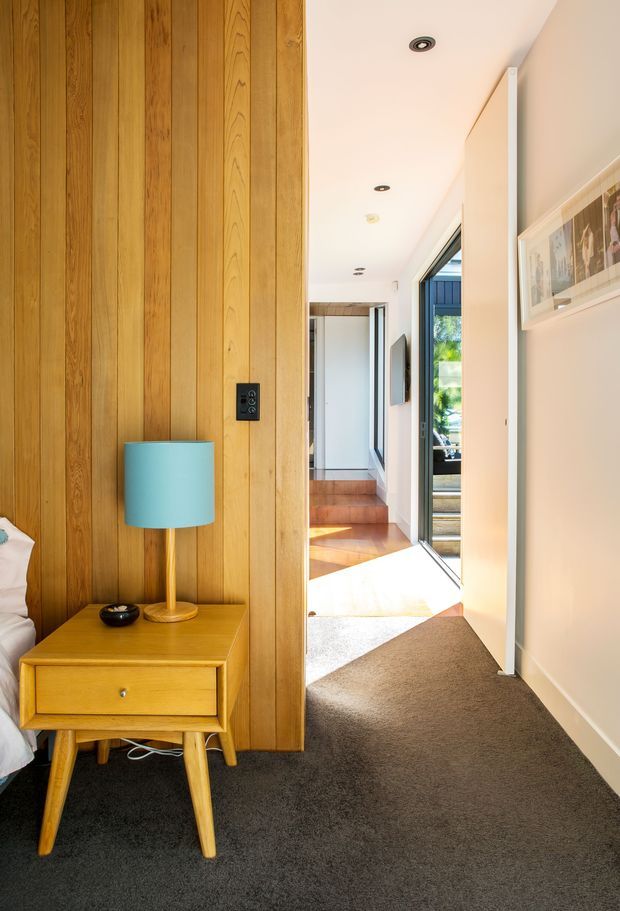 A bright residential hallway viewed from beside a bedroom, featuring a full-height vertical timber wall panel, a mid-century-style timber bedside table with a turquoise lamp, dark carpeted flooring, white walls, recessed ceiling lights, and a glazed doorway opening toward a sunlit timber staircase and exterior view.