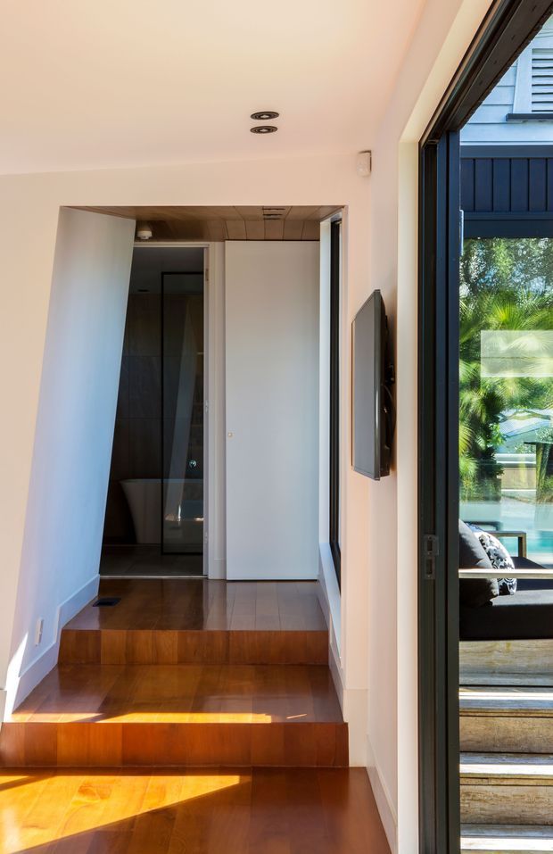 A narrow, bright interior hallway or entry passage with polished warm timber flooring and two steps leading up to a recessed bathroom area. White walls and ceiling create a clean contemporary shell, with a wood-lined ceiling section near the doorway. A wall-mounted television or display is visible on the right, beside large dark-framed glazing that looks onto an outdoor seating area and pool. The bathroom beyond includes a glass shower enclosure and freestanding or partially visible sanitary fixture.