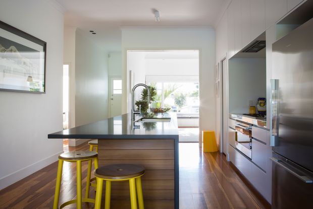 Bright contemporary galley kitchen with a central island featuring a dark stone benchtop, timber-look drawers, and two lime-yellow stools. Full-height white cabinetry and integrated appliances line the right wall, while timber flooring runs throughout. A large opening at the rear connects visually to a bright living or outdoor area, and a framed artwork hangs on the left wall.