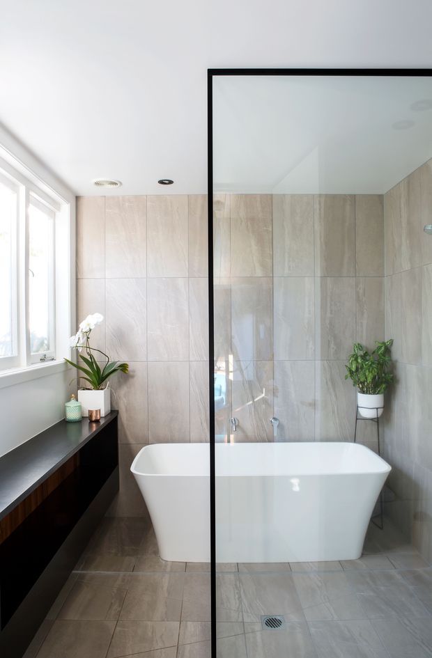 Bright contemporary bathroom with a freestanding white bathtub enclosed by a black-framed clear glass shower screen. Large pale beige-grey stone-look tiles cover the walls and floor. A dark floating vanity runs beneath the window on the left, with potted plants adding greenery. The room has clean lines, recessed ceiling lights, and abundant natural light.