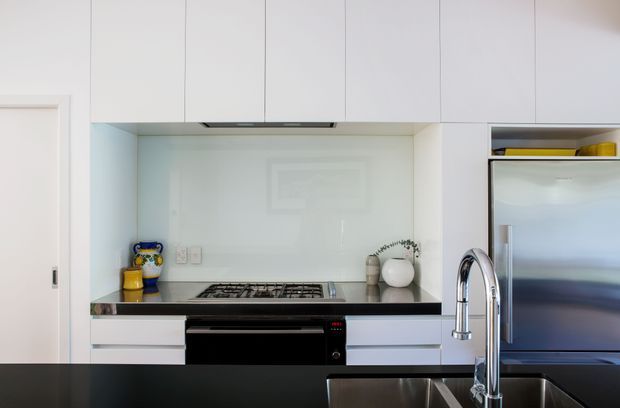 A bright, compact contemporary kitchen with white flat-panel cabinetry extending to the ceiling, a dark stone benchtop, glossy pale splashback, stainless-steel gas cooktop and oven, and a large stainless-steel refrigerator. A deep stainless-steel sink with a tall curved chrome tap occupies the foreground island or bench. Small decorative objects and pantry items add limited colour against the predominantly white and dark-grey palette.