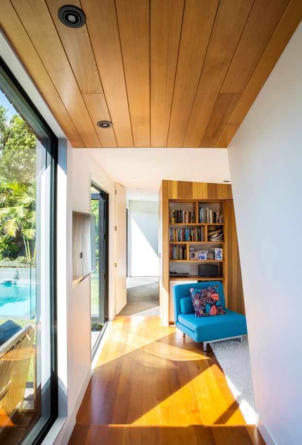 A bright contemporary interior hallway or landing with full-height glazing on the left, a warm timber-lined ceiling, polished timber flooring, white walls, and a built-in timber bookshelf. A vivid blue lounge chair with a patterned cushion sits beside the shelving, while sunlight creates strong geometric patches across the floor. Lush planting and a swimming pool are visible outside through the glazing.