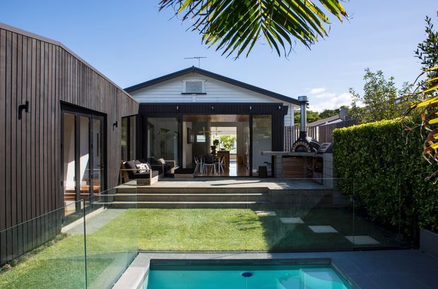 A contemporary single-storey home viewed from the rear garden, with dark vertical timber-clad wings framing a central gabled pavilion. Large glazed doors open onto a raised deck and outdoor entertaining area with a built-in barbecue, while steps lead down through a small lawn to a partially visible swimming pool. Glass balustrades, clipped hedging and tropical planting complete the landscaped courtyard-like setting.