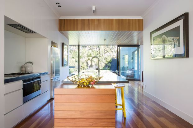 Bright contemporary kitchen viewed toward a dining and glazed living area. The room features a large timber island with a pale benchtop, streamlined white cabinetry, integrated stainless-steel appliances, dark polished timber flooring, and a timber-clad bulkhead. Full-height glazing at the rear opens the kitchen to a leafy outdoor setting, while a yellow bar stool and framed artwork add small accents.
