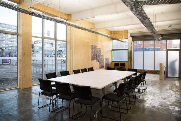Bright open-plan commercial meeting or boardroom space with a long white conference table surrounded by black chairs. The room features exposed concrete flooring, extensive timber plywood wall panelling and columns, large industrial-style windows, suspended cable trays and services, and a glazed entry door. A wall-mounted display and artwork are visible at the far end.