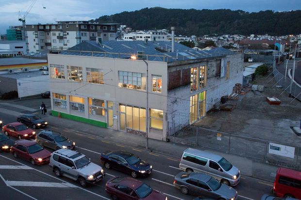 Elevated exterior view of a large two-storey industrial or commercial building on a dense urban street corner. The building has a painted light facade, extensive industrial glazing, a low pitched metal roof with roof glazing or skylight elements, and sections of exposed masonry. Adjacent apartment buildings, construction materials, hills, and traffic are visible in the surroundings.