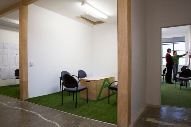 An open-plan office interior with small meeting areas separated by exposed timber posts and beams. The space has white walls, green carpeted zones, polished concrete circulation flooring, simple black-framed chairs, and a compact wood-toned meeting table with a green geometric graphic. A doorway reveals an adjoining office area with additional seating and people.