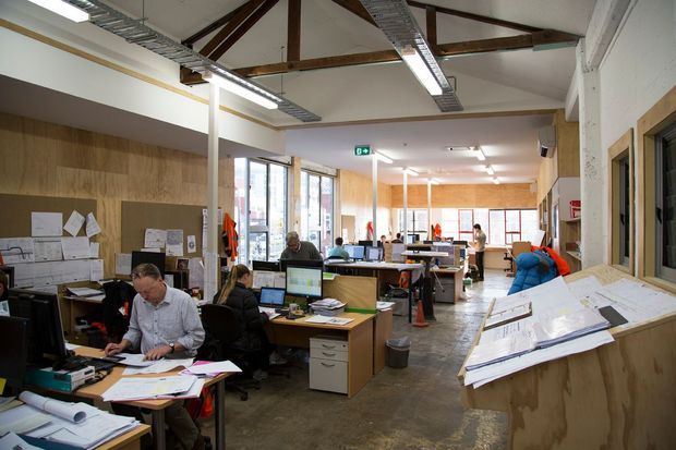 Open-plan commercial design studio or architectural office with multiple workstations, drafting tables, computers, pinned drawings, and large-format plans. The space has an industrial character with polished concrete flooring, plywood wall lining and counters, exposed timber roof trusses, high ceilings, and linear fluorescent lighting. Large windows provide abundant daylight, while staff work throughout the office.