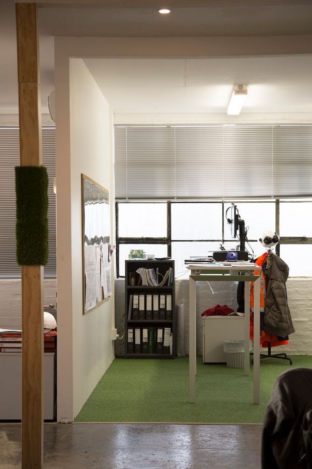A compact industrial-style office workspace viewed through a wide opening. The room has exposed timber posts, concrete flooring outside the office, green carpet, a simple metal-framed desk, office chair, filing shelves, pinned wall displays, large factory-style steel-framed windows with horizontal blinds, and exposed ceiling services with fluorescent lighting. The space is functional, bright, and utilitarian.