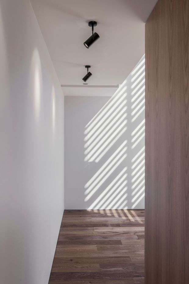 Minimalist narrow interior passage or hallway with smooth white walls and ceiling, dark cylindrical black track spotlights, warm timber plank flooring, and a tall timber-clad wall or built-in joinery element on the right. Strong diagonal bands of sunlight and shadow create a graphic feature across the far wall and floor.