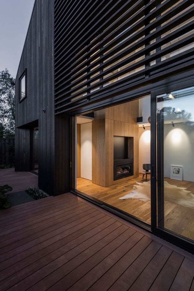 Twilight exterior view of a contemporary multi-level residence opening onto a broad timber deck. The facade combines dark vertical timber cladding with a prominent horizontal slatted privacy screen and expansive black-framed glazing. Through the open sliding doors, a warm living room is visible with timber flooring, a built-in timber fireplace wall, and a modern inset firebox.