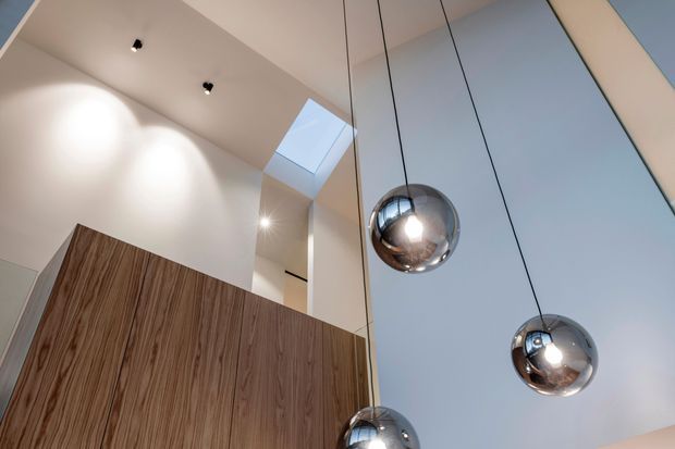 Upward-looking view of a contemporary interior with a tall white ceiling and wall surfaces, a rectangular skylight, recessed spotlights, and three suspended chrome spherical pendant lights. A large fixed timber-grain joinery or partition element occupies the lower left, while the right side appears to contain tall pale glazing or a smooth vertical wall plane.