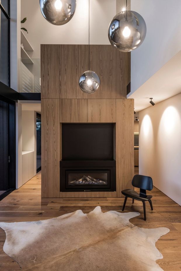A contemporary living room with a dramatic double-height volume, warm timber flooring and a full-height timber-clad fireplace wall. The fireplace surround incorporates a black firebox and television recess. Three reflective spherical pendant lights hang at different heights, while a sculptural black lounge chair and hide rug add furnishings to the room. Dark-framed glazing and built-in white shelving are visible at the sides.