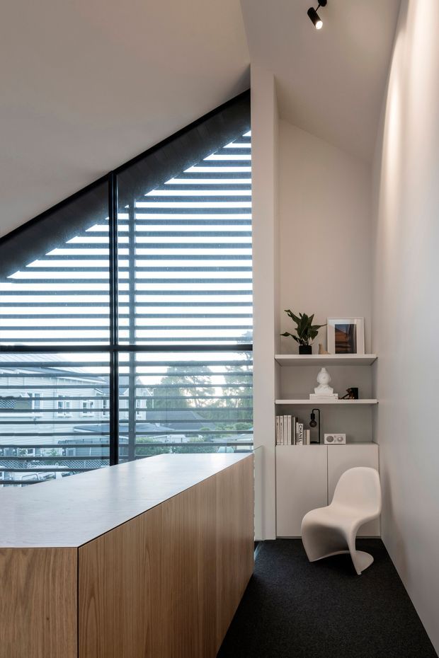 A compact, contemporary office or study nook beneath a sharply sloping ceiling. A large triangular glazed window with horizontal blinds fills the left side, while a light timber built-in desk or cabinet runs across the foreground. The right wall contains recessed open shelving and closed white cabinetry, decorated with a plant, framed artwork and small objects, alongside a sculptural white chair. Dark carpet, white walls and ceiling, and integrated spot lighting create a refined, minimalist workspace.