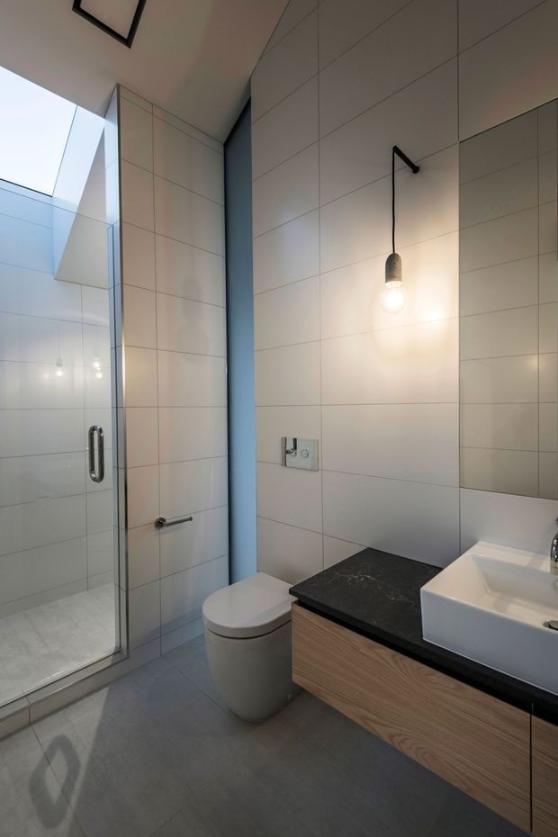 Contemporary bathroom featuring a frameless glass shower enclosure, wall-hung style toilet, floating timber-look vanity with a dark stone benchtop, white vessel basin, large pale wall tiles, grey floor tiles, and a black pendant light. A large mirror and skylight enhance the bright, clean space.