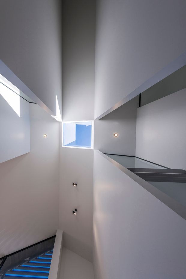 Vertical view through a modern interior stairwell or atrium, featuring sharply angled white and pale-grey walls, a square skylight/opening showing blue sky, recessed spot lighting, glass balustrades, and a dark stair flight with blue-lit treads. The composition emphasizes dramatic geometry, verticality, and abundant natural light.