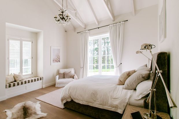 Bright, elegant bedroom with a large upholstered bed, layered neutral bedding, white walls, exposed painted ceiling beams, tall curtained windows, plantation shutters, a built-in window seat, chandelier, and a vintage-style floor lamp. The room has warm timber flooring and a soft, refined neutral palette.