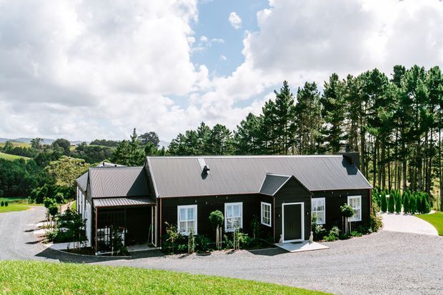 A single-storey rural house with dark vertical cladding, a long corrugated pitched roof, white-trimmed windows and doors, and a covered entry porch. The home sits in a landscaped countryside setting with gravel driveways, lawns, mature trees and rolling farmland beyond.