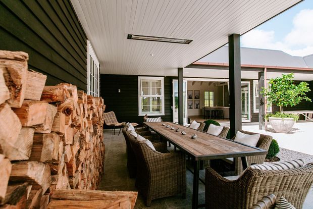 A covered outdoor dining terrace with a long timber dining table surrounded by woven wicker chairs. The space features a white-lined ceiling, dark horizontal cladding, black structural posts, a stacked firewood feature, and views into a bright courtyard with planted pots and adjoining buildings.
