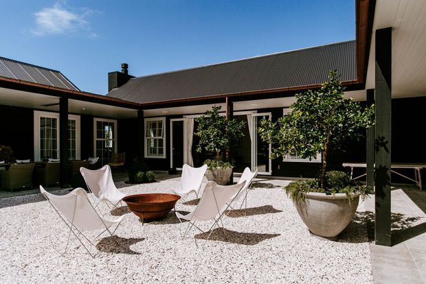 A bright landscaped courtyard enclosed by a dark-clad, single-storey building with pitched corrugated metal roofs, white-framed windows and doors, and covered verandah-like edges. A white gravel surface forms an outdoor seating area with sculptural sling chairs, a central fire bowl, and large potted trees and plants.