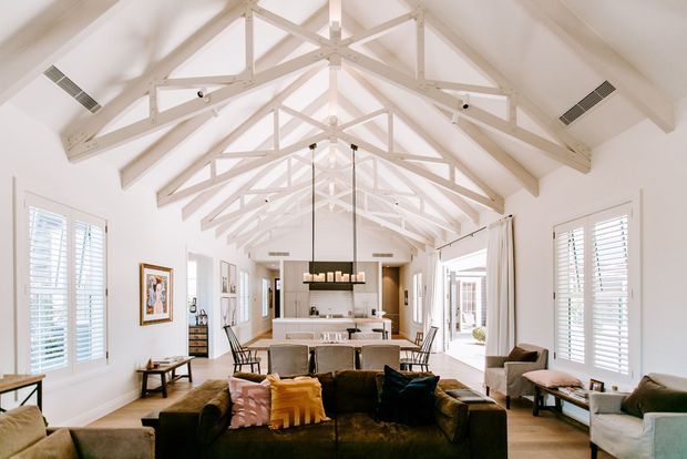 Bright open-plan living room with a dramatic vaulted ceiling, exposed painted timber trusses and beams, white plantation shutters, and extensive glazing. A large dark sectional sofa faces a contemporary kitchen and dining area, with a long linear pendant light above the island. The space has a relaxed yet refined coastal character and strong indoor-outdoor connection.