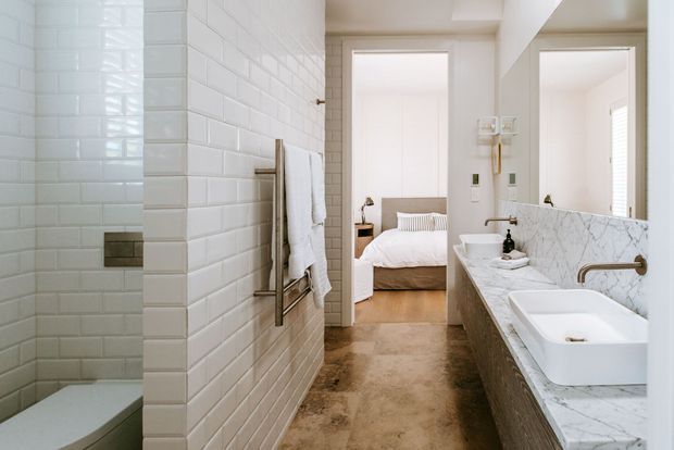 Bright, refined bathroom with a walk-in shower enclosed in glossy white subway tiles, a freestanding-style bathtub edge in the foreground, and a long marble-look vanity with a white vessel basin and brass tapware. A bedroom is visible through the open doorway, with warm stone-toned flooring continuing through the bathroom and a large mirror above the vanity.
