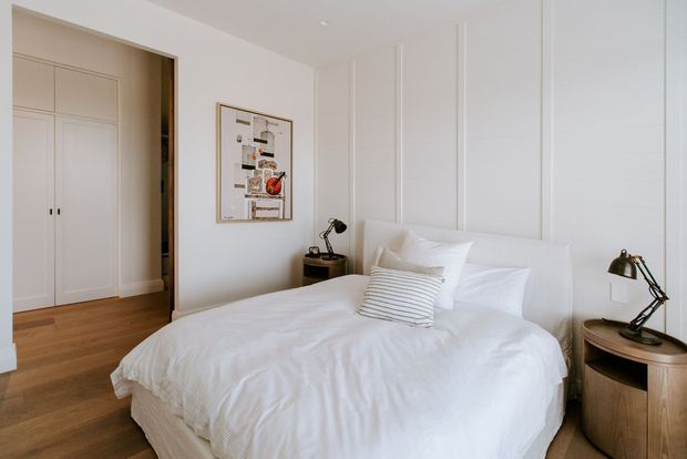 Bright, refined bedroom with a white upholstered bed, layered white bedding, and a striped accent cushion. The room features warm timber flooring, white paneled walls, integrated white wardrobe cabinetry, matching dark metal bedside lamps, and rounded timber bedside tables. A framed artwork adds a small visual accent.