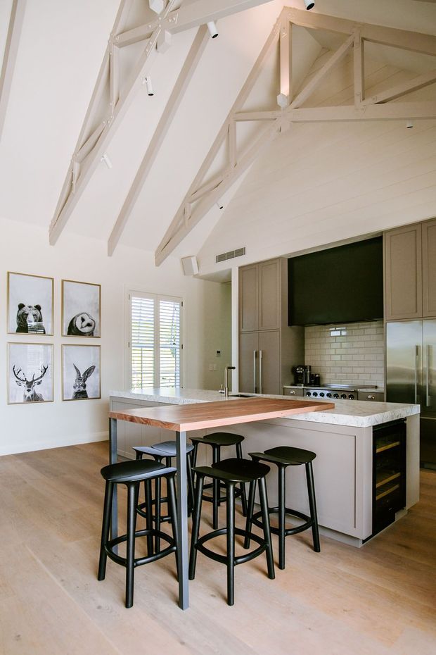 Bright, open-plan kitchen with a large island featuring a white stone benchtop, integrated timber breakfast bar, and black bar stools. The kitchen has taupe-grey shaker cabinetry, stainless-steel appliances, a white subway-tile splashback, pale timber flooring, and a dramatic vaulted ceiling with exposed painted timber trusses and integrated spotlights. Plantation shutters and framed artwork add detail to the adjacent wall.