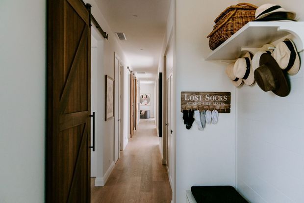 A bright residential hallway with warm timber flooring, white walls, multiple doorways, and a large dark-stained sliding barn door in the foreground. Built-in white shelving displays woven baskets and hats, while a wall-mounted lost-socks organizer and a small black bench add practical storage. The corridor extends toward a softly furnished room in the distance.