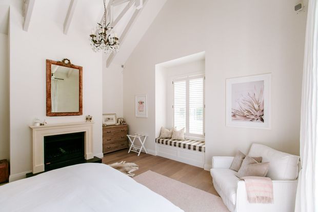Bright, elegant bedroom with a high vaulted ceiling, exposed painted beams, a decorative chandelier, white fireplace mantel with dark insert, timber-framed mirror, built-in window seat with plantation shutters, dresser, upholstered sofa, and a soft neutral palette. The room combines classic architectural detailing with refined coastal-inspired furnishings.