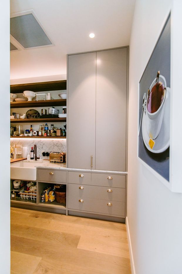 A compact, refined kitchen or butler's pantry with full-height pale grey cabinetry, open dark timber shelving, a marble-patterned splashback, integrated drawers, and a white farmhouse-style sink. The space has warm timber flooring, under-shelf lighting, recessed ceiling lighting, and a framed coffee-themed artwork on the wall.
