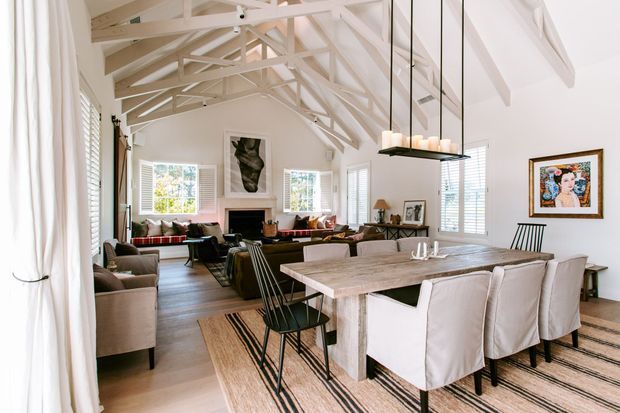 Bright open-plan interior combining a dining area and living room beneath a high pitched ceiling with exposed painted timber trusses and beams. The space features white walls, extensive white plantation shutters, pale timber flooring, a large rustic timber dining table with upholstered dining chairs, a linear suspended candle-style light, and a relaxed seating area around a fireplace. Artwork and layered rugs add visual warmth.