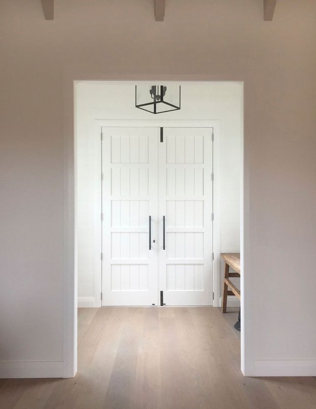 A bright, restrained interior view focused on a pair of white paneled double doors with dark vertical hardware and exposed hinges. The doors sit within a wide white-trimmed opening, with pale timber flooring, soft white walls, a simple black geometric pendant light, and partial timber furniture visible beyond the doorway. Exposed ceiling beams add subtle architectural detail.