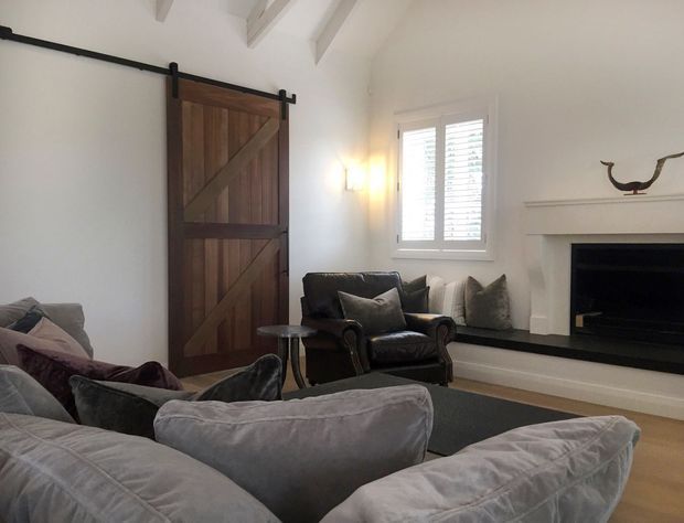 A cozy living room with a large sectional sofa, leather armchair, dark rug, white walls, exposed white ceiling beams, a rustic timber barn-style sliding door, shuttered window, and a white fireplace surround with a dark hearth and firebox. Neutral furnishings and warm wall lighting create a relaxed, refined atmosphere.