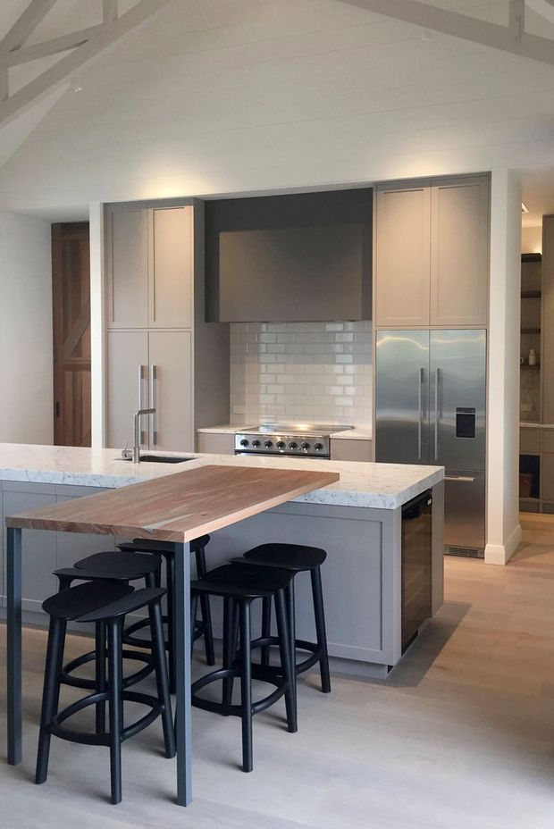 A bright, refined kitchen with a large central island featuring pale stone benchtops, grey shaker-style cabinetry, a timber breakfast bar extension, and black counter stools. The rear wall includes integrated tall cabinetry, a stainless-steel refrigerator and range, a dark feature recess, and a white subway-tile splashback. White-painted exposed roof beams and light timber flooring contribute to an airy, contemporary farmhouse character.