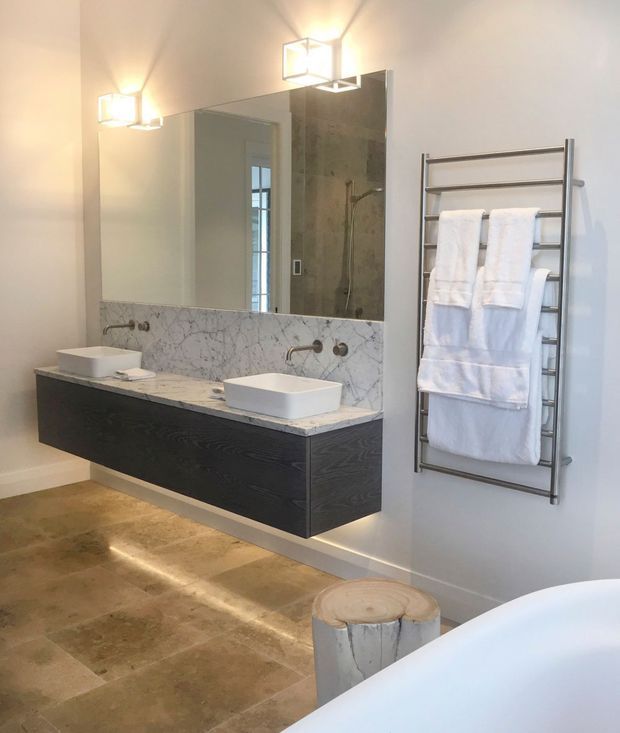 Bright contemporary bathroom featuring a floating dark timber double vanity with white vessel basins, a veined marble backsplash, large wall mirror, chrome heated towel rail, stone-look floor tiles, and a partially visible freestanding bathtub. Warm integrated lighting beneath the vanity and geometric wall sconces add a refined ambience.