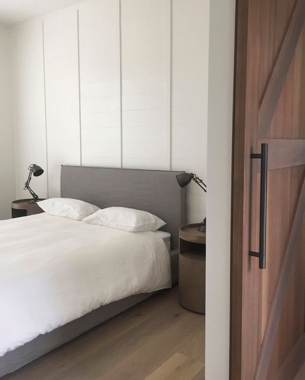 A compact, refined bedroom viewed through an open timber door. The room features a low upholstered grey bedhead, white bedding, matching dark bedside tables with black articulated lamps, pale timber flooring, and a white vertically panelled feature wall. The door has rich natural timber framing and a long black vertical handle.