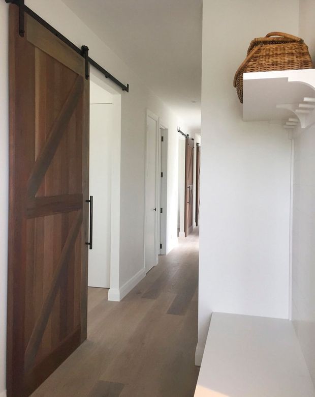 Bright residential hallway with white walls, pale timber flooring, multiple white internal doors, and dark timber barn-style sliding doors on black metal hardware. A built-in white bench and curved wall shelf create a small entry or hallway nook, with a woven basket stored above.
