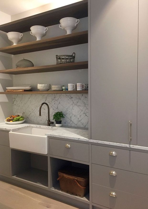 A refined kitchen or butler's pantry with matte grey built-in cabinetry, open dark timber shelves displaying crockery, a white farmhouse sink, aged brass or bronze tapware and hardware, and a veined marble backsplash and benchtop. The space has warm, softly lit, traditional-meets-contemporary detailing.