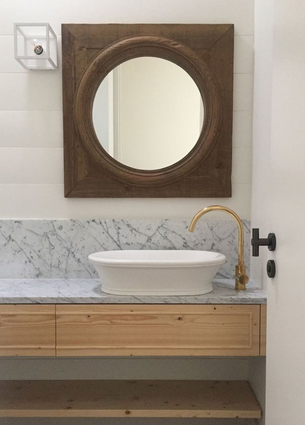 A refined bathroom vanity featuring a white vessel basin on a light timber floating cabinet, a white-and-grey marble backsplash and benchtop, and a brass wall-mounted curved tap with black controls. A large round mirror sits within a dark timber square frame above the vanity, complemented by a small wall light.