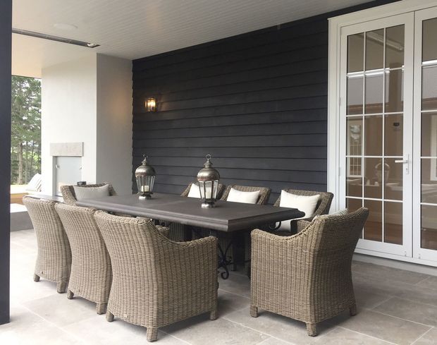 Covered outdoor dining terrace with a large dark dining table surrounded by woven wicker armchairs. The space features a dark horizontal-clad feature wall, white-framed glazed French doors, pale tiled flooring, lantern-style lighting, and a bright open side overlooking greenery.