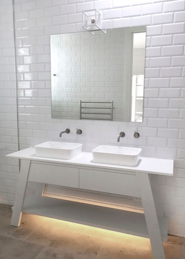 Bright contemporary bathroom featuring a double vanity with two white vessel basins, wall-mounted brushed metal tapware, a large frameless mirror, white glossy brick-pattern wall tiles, and an illuminated open vanity base.